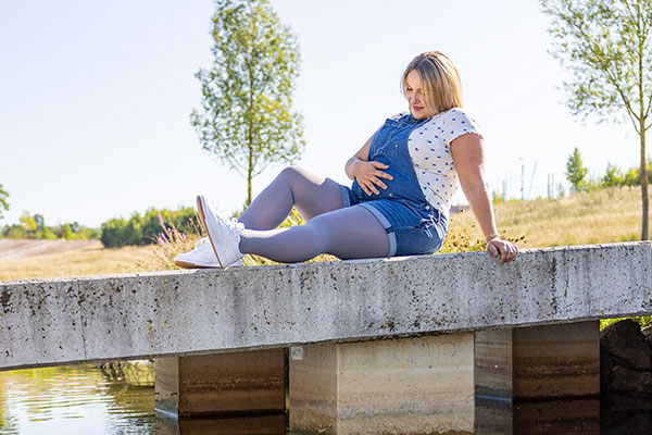 Eine schwangere Frau sitzt auf einer Brücke und streichelt ihren Babybauch.